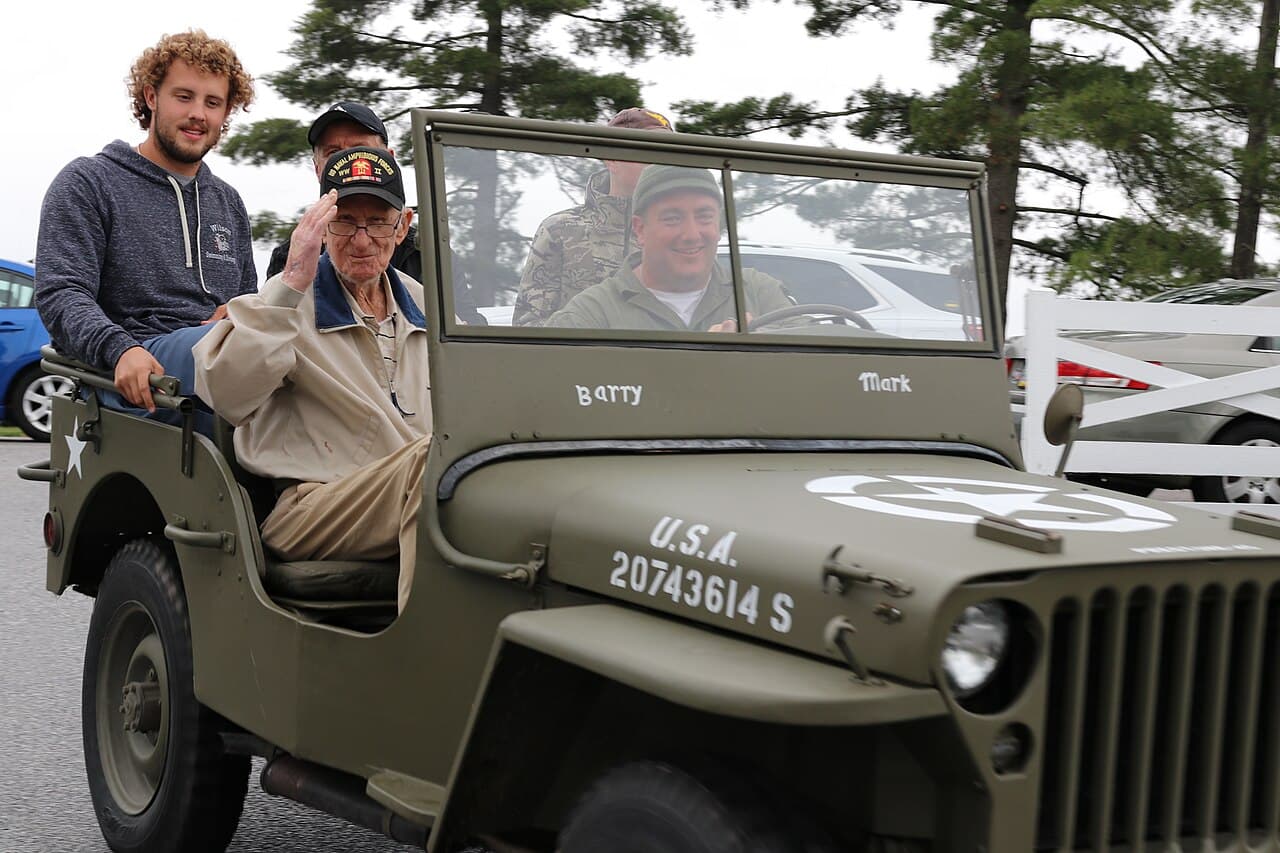 A man in a ballcap and jacket sits in the front seat of a Jeep, saluting the camera, with others seated behind him.…