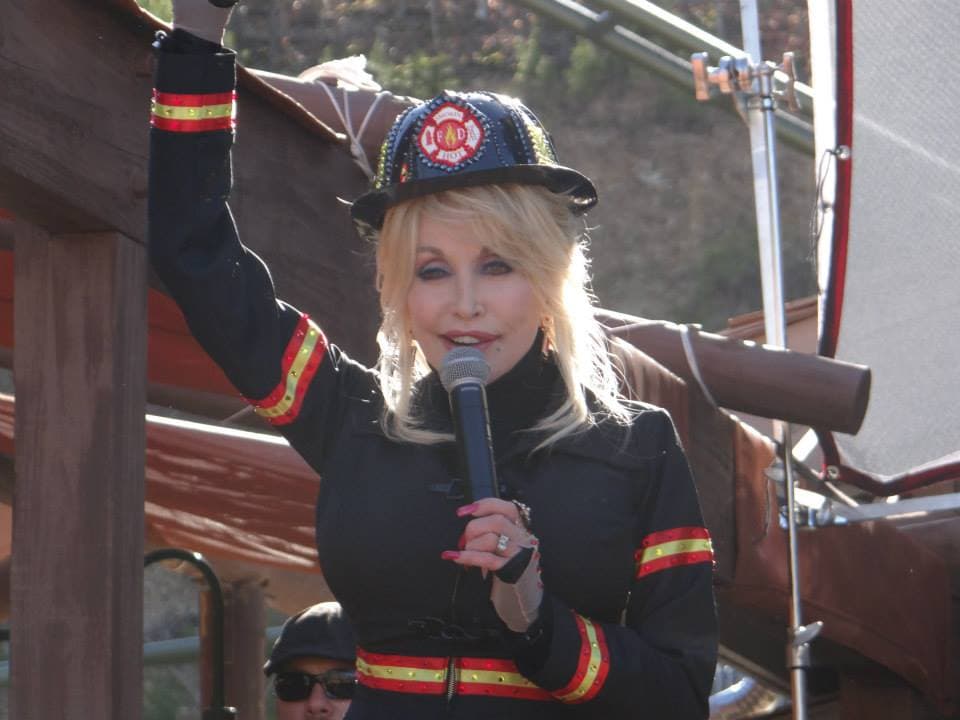 Dolly Parton, vestida con ropa de bombero, en Dollywood, presentando nuevas atracciones. Tiene un micrófono en sus…